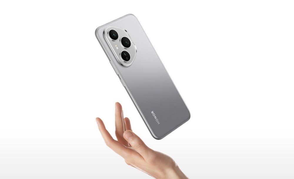 Na obrazie widoczny jest szary smartfon z trzema aparatami na tylnej obudowie, unoszący się nad dłonią. Na obudowie znajduje się napis \'HONOR\'.