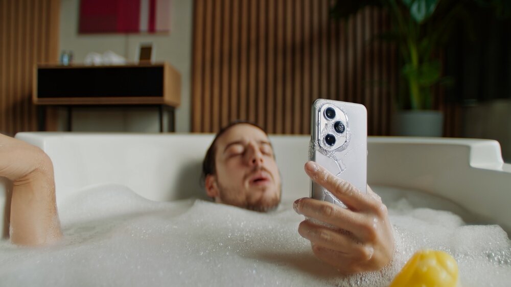 Osoba leży w wannie pełnej piany, trzymając w ręku smartfon z trzema widocznymi obiektywami aparatu. W tle znajduje się drewniana ściana i roślina doniczkowa.