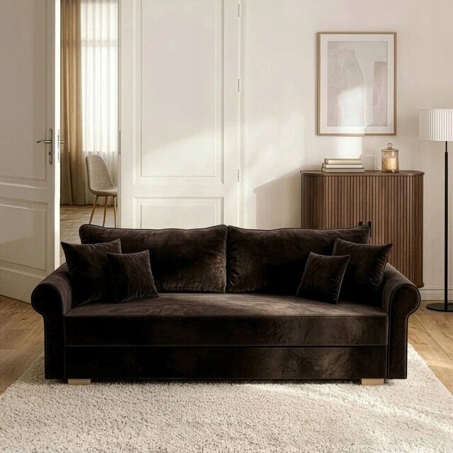Brązowa sofa rozkładana z funkcją spania, ustawiona na jasnym dywanie w eleganckim salonie. W tle znajduje się drewniana komoda z książkami i dekoracjami oraz stojąca lampa.