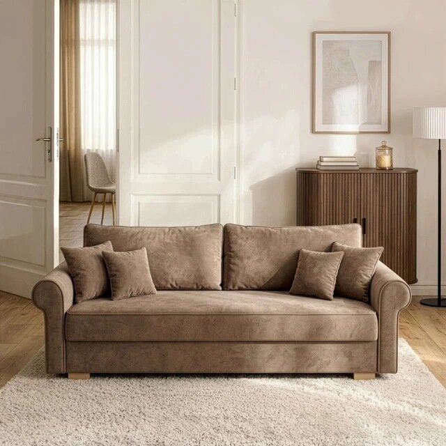 Beżowa sofa rozkładana z funkcją spania, otoczona kilkoma poduszkami, stoi na jasnym dywanie w eleganckim salonie. W tle znajduje się drewniana komoda z książkami i dekoracyjnym wazonem oraz stojąca lampa.
