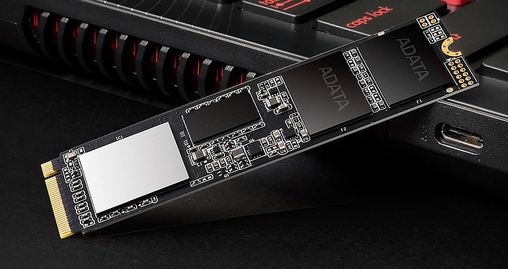 Na obrazie widoczny jest dysk SSD marki ADATA umieszczony na tle klawiatury laptopa. Dysk ma czarną obudowę z widocznymi elementami elektronicznymi i złączem M.2.
