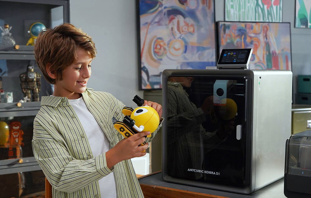 Osoba trzyma żółty obiekt przypominający robota przed drukarką 3D z napisem \'ANYCUBIC KOBRA S1\'. W tle widoczne są półki z różnymi przedmiotami i kolorowe obrazy na ścianie.