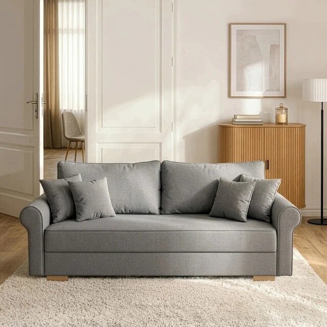 Szara sofa rozkładana z funkcją spania, otoczona kilkoma poduszkami, stoi na jasnym dywanie w eleganckim salonie. W tle znajduje się drewniana szafka z książkami i dekoracjami oraz lampa stojąca.
