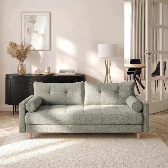 Szara sofa rozkładana z funkcją spania stoi na jasnym dywanie w eleganckim salonie. W tle znajduje się ciemna komoda z dekoracjami oraz lampa stojąca.