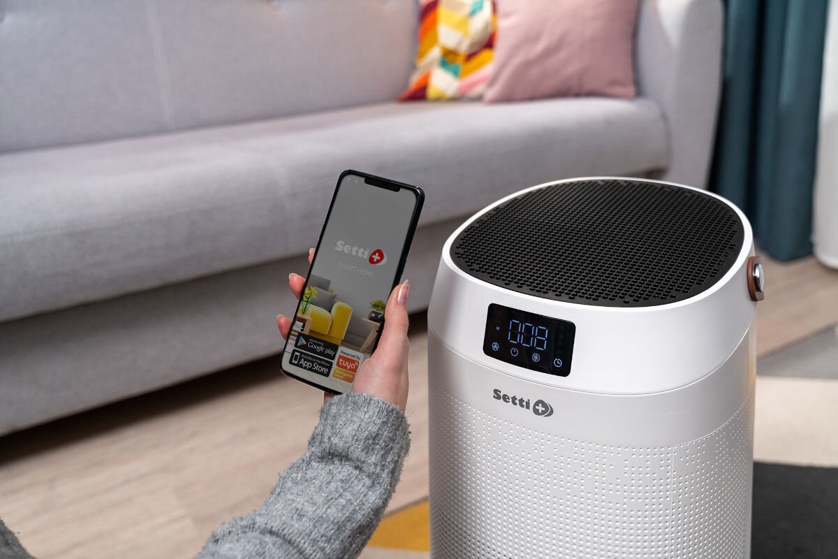 Na obrazie widoczny jest biały oczyszczacz powietrza z wyświetlaczem cyfrowym i logo \'Setti+\'. Obok osoba trzyma smartfon z aplikacją \'Setti+\' na ekranie.