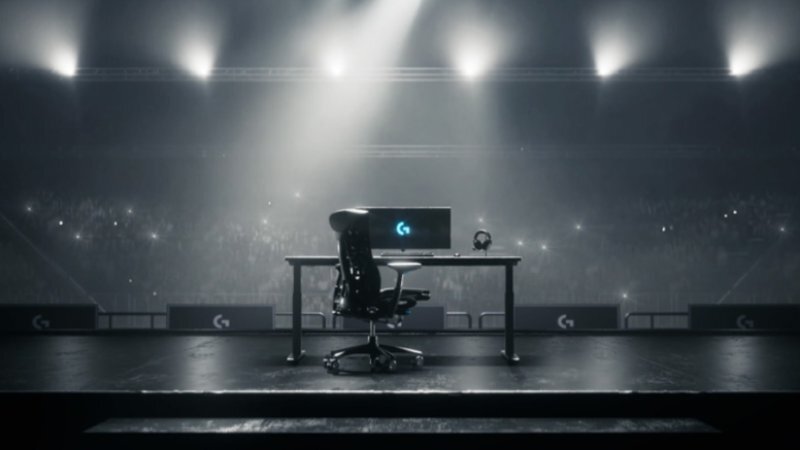 Na scenie znajduje się biurko z monitorem, krzesłem i słuchawkami, a w tle widać rozświetlone reflektorami trybuny z publicznością. Na monitorze widoczne jest logo \'G\'.