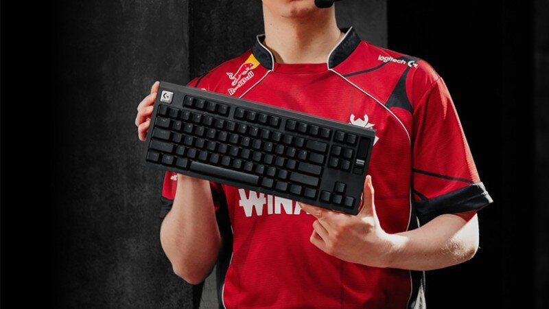 Osoba w czerwonej koszulce trzyma czarną klawiaturę mechaniczną. Na koszulce widoczne są logotypy, w tym \'logitech\' i \'Red Bull\'.