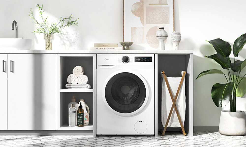 PRALKA MIDEA MF110W80BA10/WW-PL Pralka Midea zabudowana pod blatem w jasnej pralni lub łazience. Grafika prezentuje urządzenie w otoczeniu półek na ręczniki i środki do prania