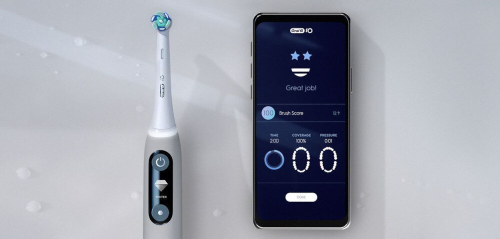 Na obrazie widoczna jest szara szczoteczka magnetyczna z logo ORAL-B oraz smartfon z aplikacją pokazującą wyniki szczotkowania. Na ekranie telefonu widnieje tekst: \'Great job!\', \'Brush Score\', \'TIME 2:00\', \'COVERAGE 100%\', \'PRESSURE 001\'.
