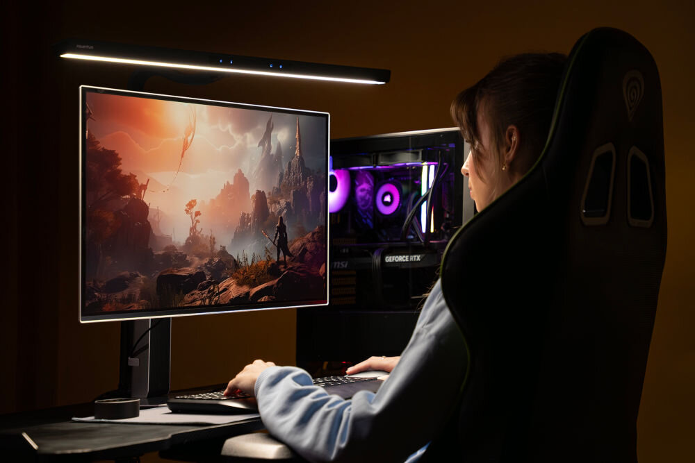 Osoba siedzi przy biurku, patrząc na monitor z wyświetloną sceną z gry fantasy. Nad monitorem zamontowana jest lampka, a w tle widać komputer z podświetleniem RGB.