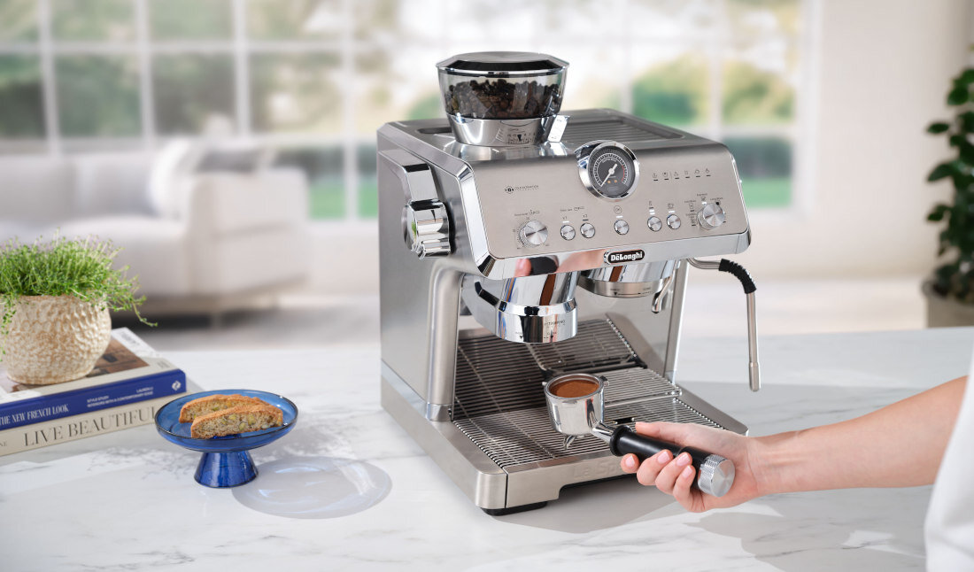 Srebrny ekspres do kawy z logo \'DeLonghi\' stoi na blacie, obok filiżanki z kawą. W tle widać rozmyty salon z rośliną i książką na stole.