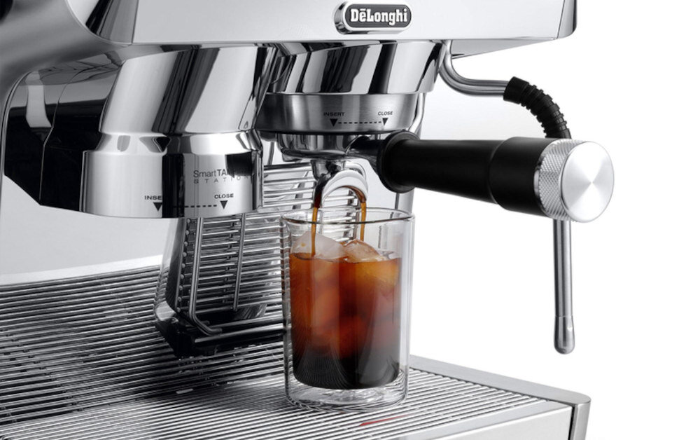 Ekspres do kawy z logo \'DeLonghi\' nalewa kawę do szklanki z lodem. Urządzenie ma metaliczne wykończenie i widoczny uchwyt do parzenia kawy.