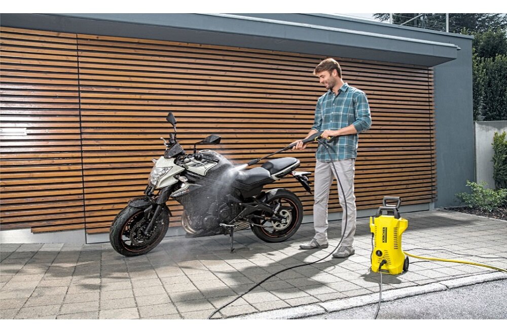 Mężczyzna używa żółtej myjki ciśnieniowej do czyszczenia motocykla na tle drewnianej ściany. Na myjce widoczne jest logo \'KARCHER\'.