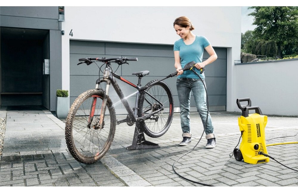 Osoba myje rower za pomocą żółtej myjki ciśnieniowej z widocznym logo KARCHER. W tle znajduje się nowoczesny budynek z szarymi drzwiami garażowymi.