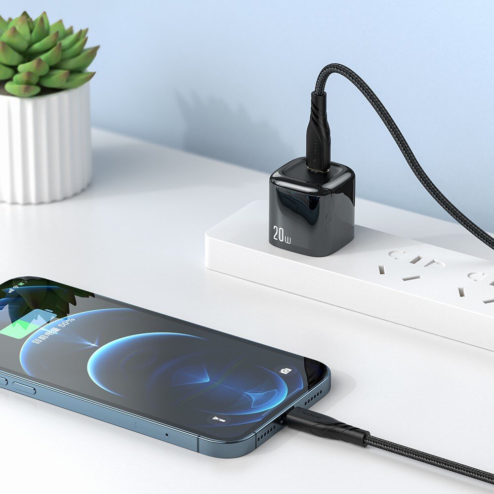 Na białym blacie znajduje się czarny adapter z napisem \'20W\' podłączony do listwy zasilającej oraz telefon z podłączonym kablem USB-C. Obok stoi mała doniczka z zieloną rośliną.