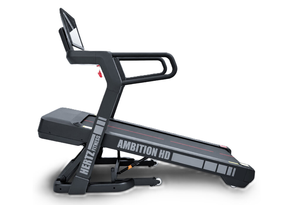 Na obrazie widoczna jest bieżnia elektryczna z napisem \'HERTZ FITNESS AMBITION HD\'. Bieżnia ma ciemny kolor z jasnymi elementami i wyświetlaczem na górze.