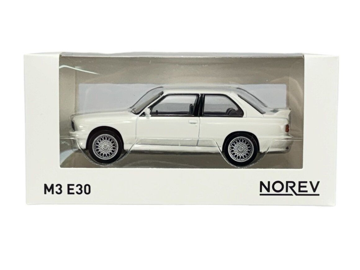 Model samochodu w skali 1:43 w kolorze białym, umieszczony w przezroczystym opakowaniu z napisem \'M3 E30\' i logo \'NOREV\'.