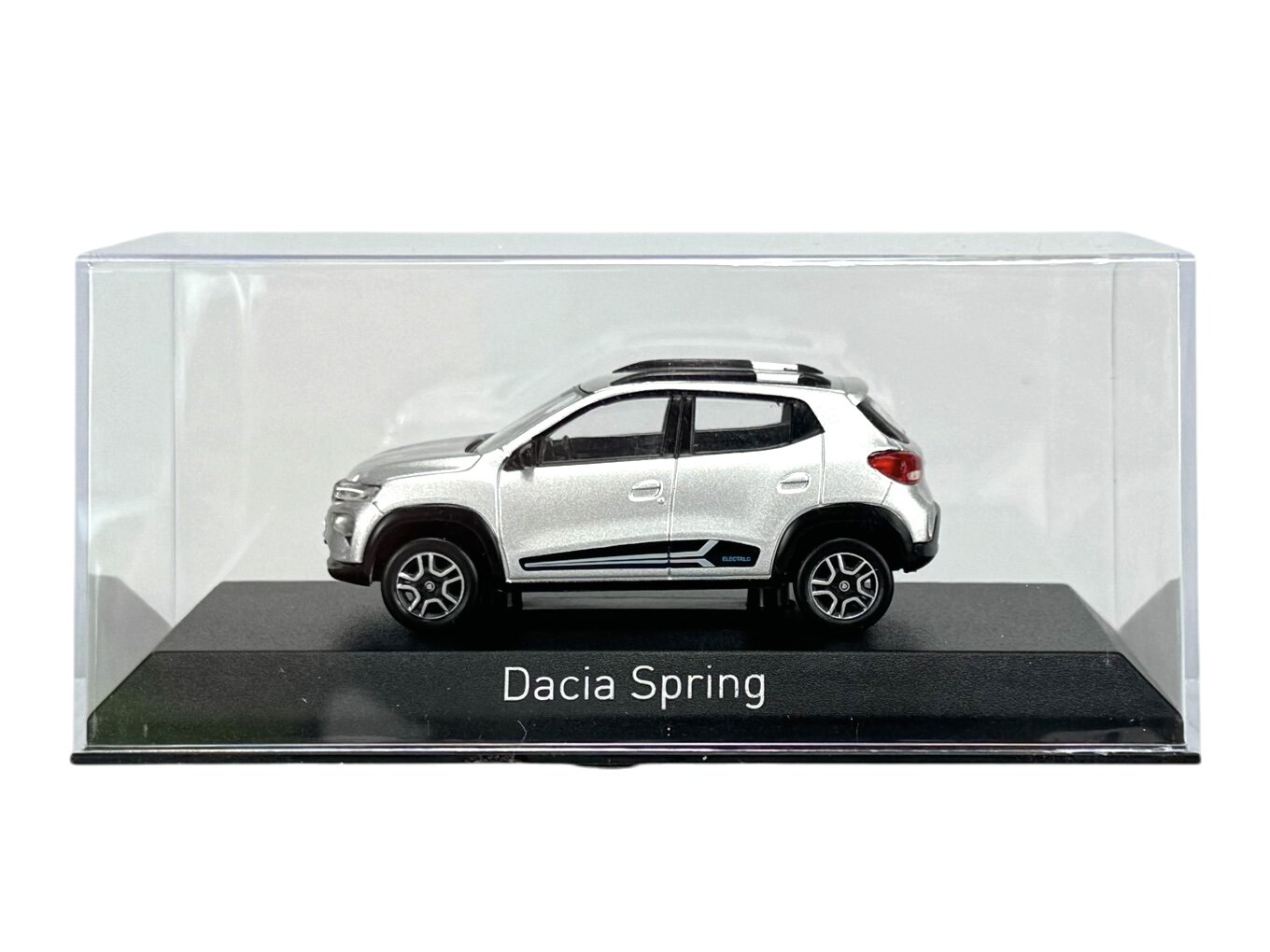 Model samochodu Dacia Spring w srebrnym kolorze, umieszczony w przezroczystej gablocie na czarnej podstawce z napisem \'Dacia Spring\'.