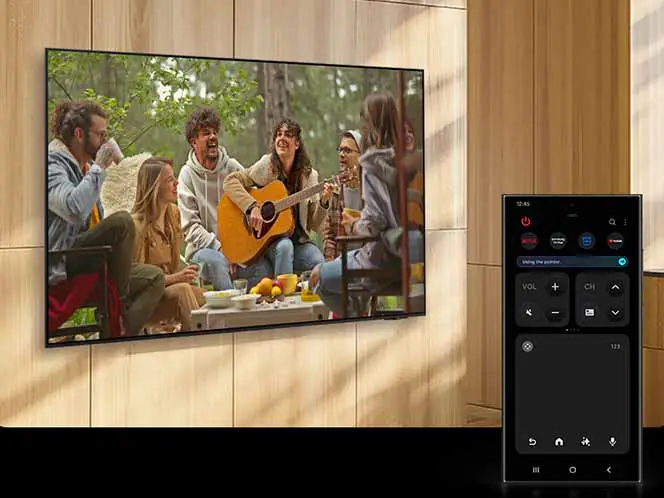 Na ścianie wisi telewizor wyświetlający obraz grupy osób siedzących na zewnątrz, z jedną osobą grającą na gitarze. Obok telewizora widoczny jest smartfon z aplikacją do zdalnego sterowania, pokazującą przyciski do regulacji głośności i zmiany kanałów.