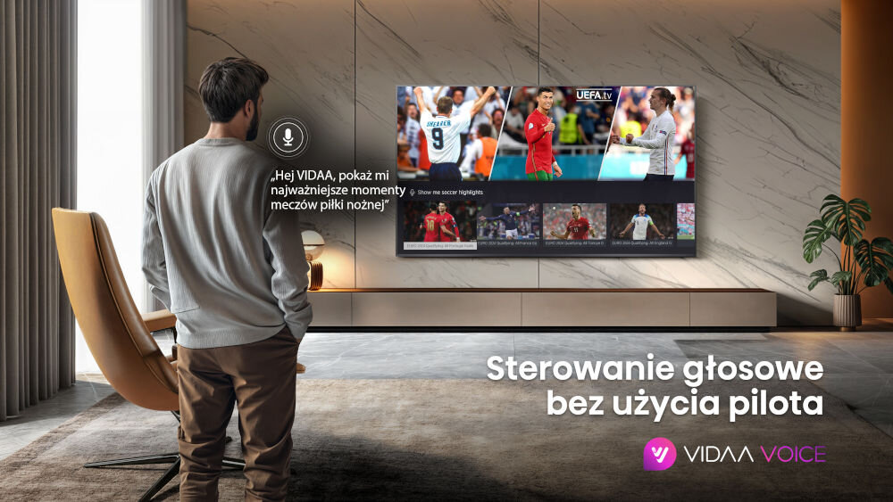 Mężczyzna stoi w salonie przed telewizorem, na którym wyświetlane są mecze piłki nożnej. Na ekranie widoczny jest tekst: \'Hej VIDAA, pokaż mi najważniejsze momenty meczów piłki nożnej\' oraz \'Sterowanie głosowe bez użycia pilota\'.