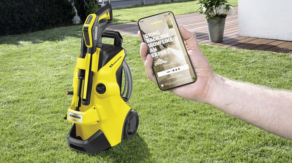 Żółta myjka ciśnieniowa z widocznym logo KARCHER stoi na trawie. Osoba trzyma smartfon z tekstem \'BRING BACK THE WOW LIKE AN EXPERT.\'.