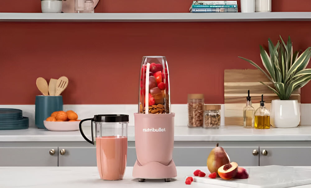 Na blacie kuchennym stoi różowy blender z logo \'nutribullet\', wypełniony owocami i orzechami. Obok znajduje się kubek z różowym napojem, a w tle widać półki z książkami i rośliną.