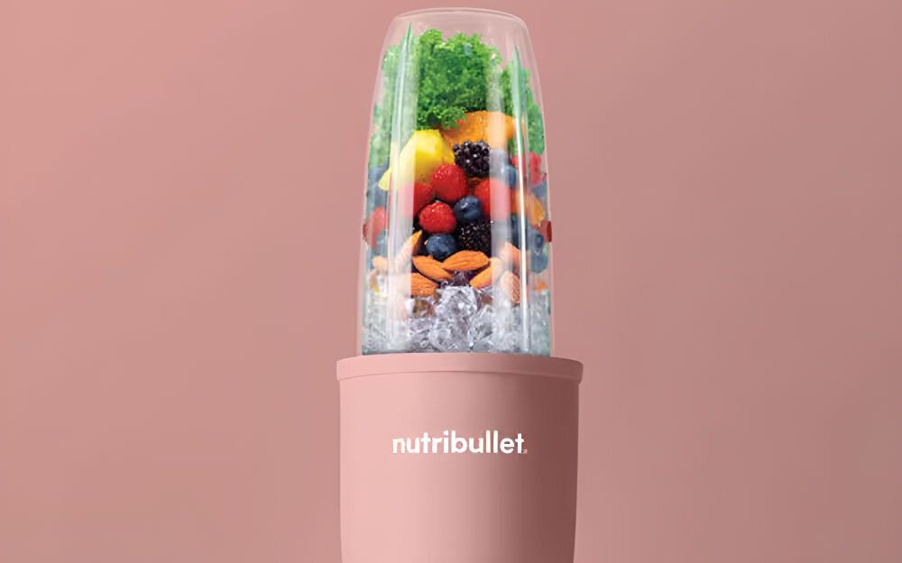 Blender personalny w kolorze różowym z logo \'nutribullet\' na obudowie. W pojemniku znajdują się różnorodne owoce, warzywa i kostki lodu.