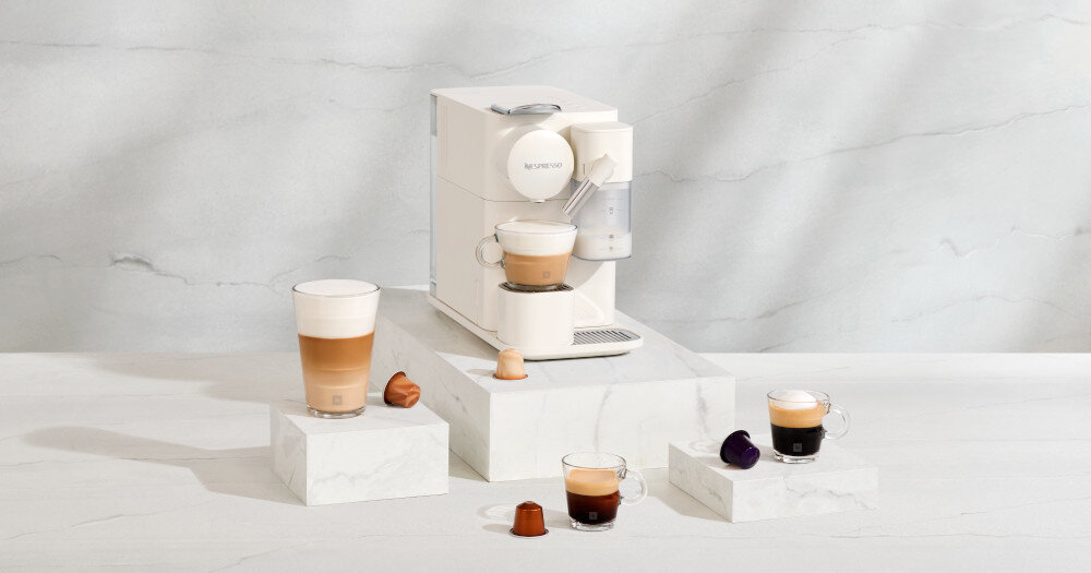 Na marmurowej powierzchni stoi biały ekspres do kawy z logo \'Nespresso\', obok którego znajdują się filiżanki z kawą i kapsułki. Ekspres ma kompaktowy design i jest otoczony różnymi rodzajami kawy w szklanych filiżankach.