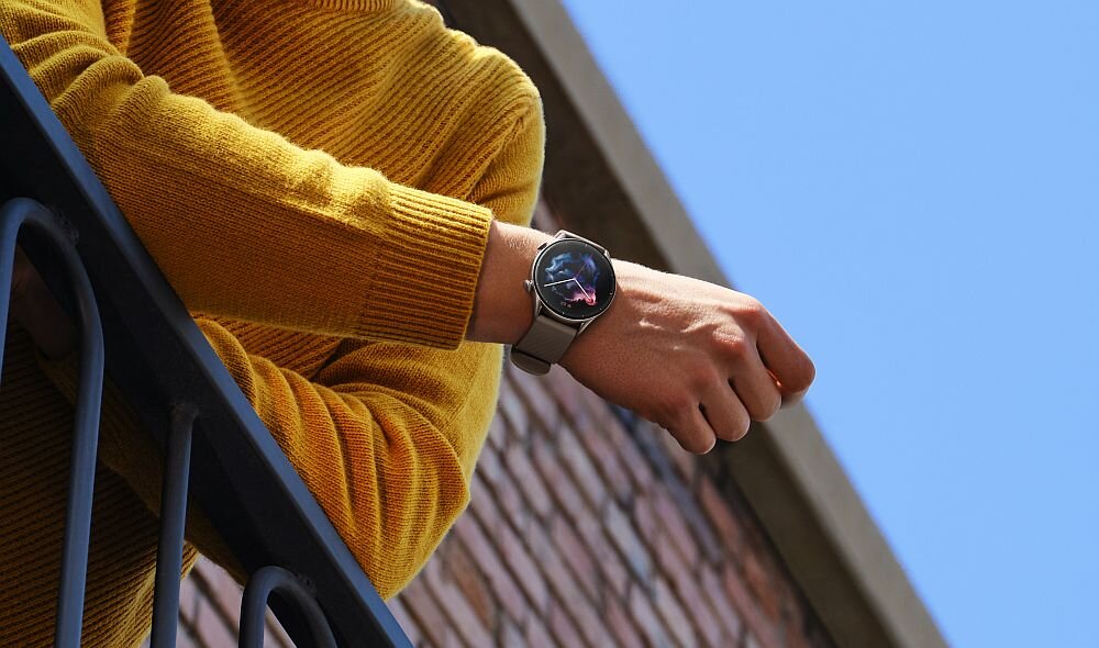 Osoba w żółtym swetrze stoi przy balustradzie, nosząc smartwatch na nadgarstku. W tle widoczna jest ceglana ściana i niebieskie niebo.