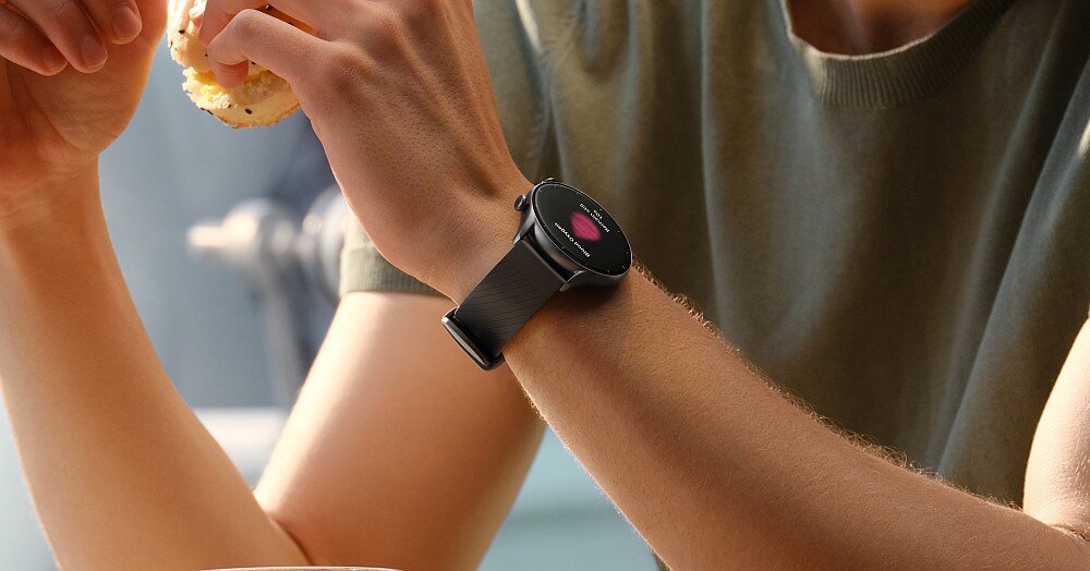 Osoba nosi smartwatch z okrągłym wyświetlaczem i ciemnym paskiem. Na ekranie zegarka widoczny jest kolorowy wskaźnik.