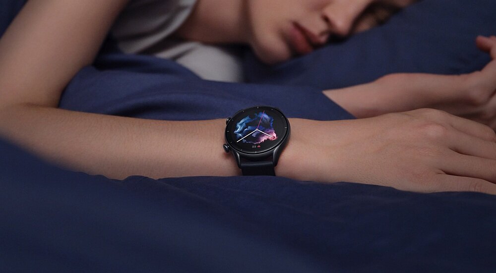 Osoba leży na łóżku, mając na nadgarstku smartwatch z okrągłym wyświetlaczem, na którym widoczna jest kolorowa grafika. Tło stanowi ciemnoniebieska pościel.