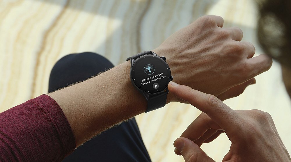 Osoba nosi smartwatch z okrągłym wyświetlaczem, na którym widoczny jest tekst: \'Measure your heart, relax more and live long.\'