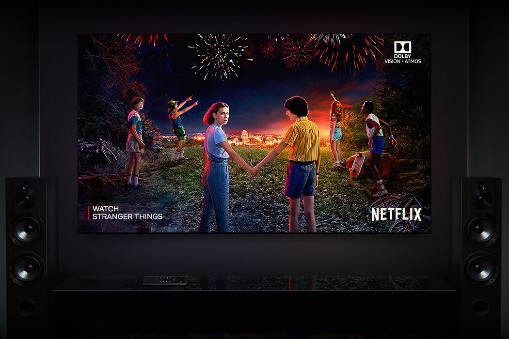 Na ekranie telewizora wyświetlany jest obraz z serialu \'Stranger Things\', przedstawiający grupę dzieci trzymających się za ręce na tle fajerwerków. Widoczne są logotypy \'Dolby Vision - Atmos\' oraz \'Netflix\'.