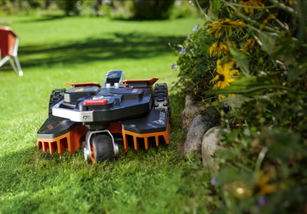 Robot koszący na trawie obok kwitnących roślin, z widocznymi elementami w kolorze czarnym i pomarańczowym. Na urządzeniu widoczny jest napis \'Landroid\'.