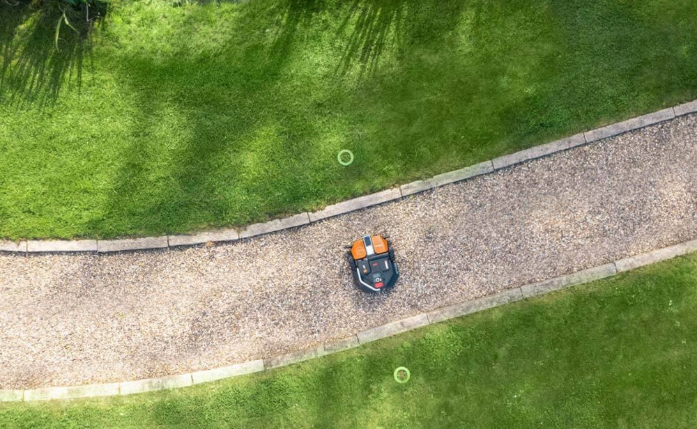 Na obrazie widać robot koszący poruszający się po żwirowej ścieżce otoczonej trawnikiem. Robot ma czarną obudowę z pomarańczowymi elementami.