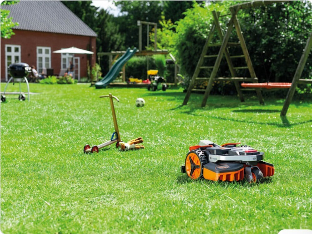 Na trawniku znajduje się robot koszący o pomarańczowych elementach. W tle widoczne są zabawki, zjeżdżalnia i huśtawka w ogrodzie przy domu.