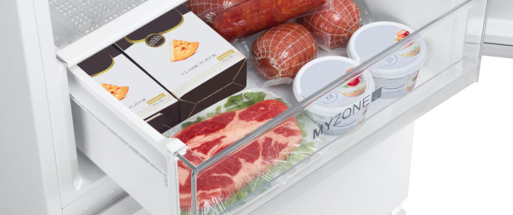 Wnętrze lodówki z szufladą zawierającą różne produkty spożywcze, w tym mięso, sery w opakowaniach, jogurty oraz wędliny. Na szufladzie widoczny jest napis \'MY ZONE\'.