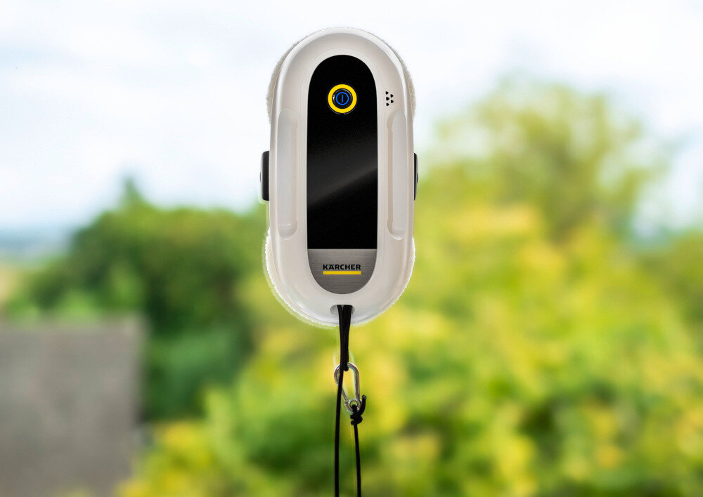 Na zdjęciu widoczny jest robot do mycia okien z logo \'KARCHER\' na przedniej części. W tle znajduje się rozmyty widok zieleni.