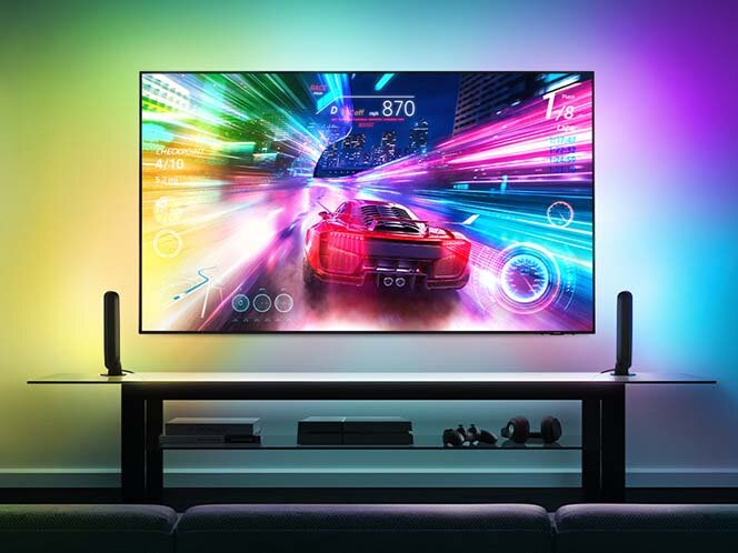 Na obrazie widoczny jest telewizor wyświetlający dynamiczną scenę z gry wyścigowej, z kolorowymi efektami świetlnymi wokół ekranu. Przed telewizorem znajduje się szafka z dwoma głośnikami po bokach.