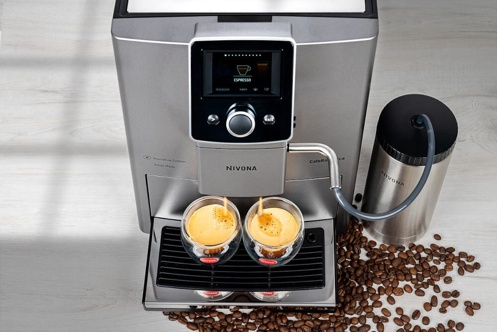 Ekspres do kawy z wyświetlaczem pokazującym napis \'ESPRESSO\', z którego do dwóch szklanek nalewana jest kawa. Obok ekspresu znajduje się pojemnik z napisem \'NIVONA\', a wokół rozsypane są ziarna kawy.