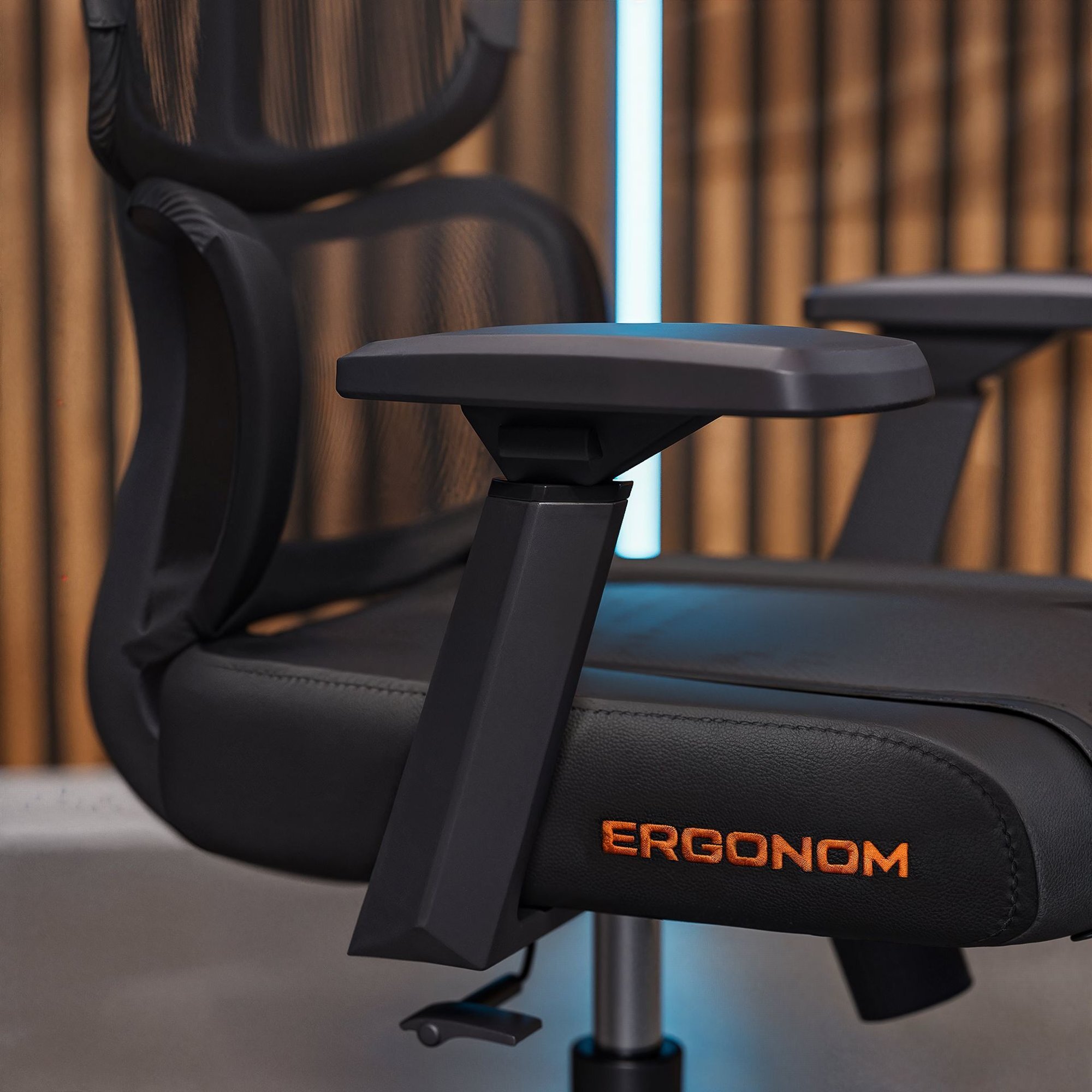 Czarny fotel z siatkowanym oparciem i regulowanym podłokietnikiem, z widocznym napisem \'ERGONOM\' na siedzisku.