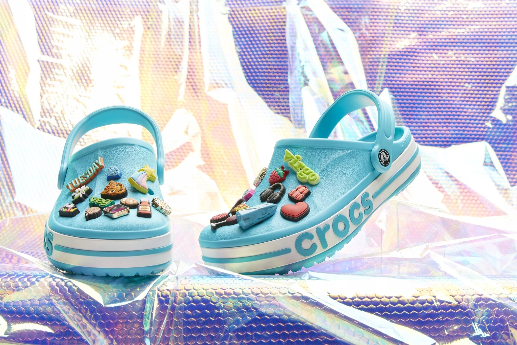 Para niebieskich chodaków z ozdobnymi przypinkami na wierzchu, z widocznym napisem \'crocs\' na boku podeszwy. Tło jest kolorowe i błyszczące, przypominające holograficzną powierzchnię.