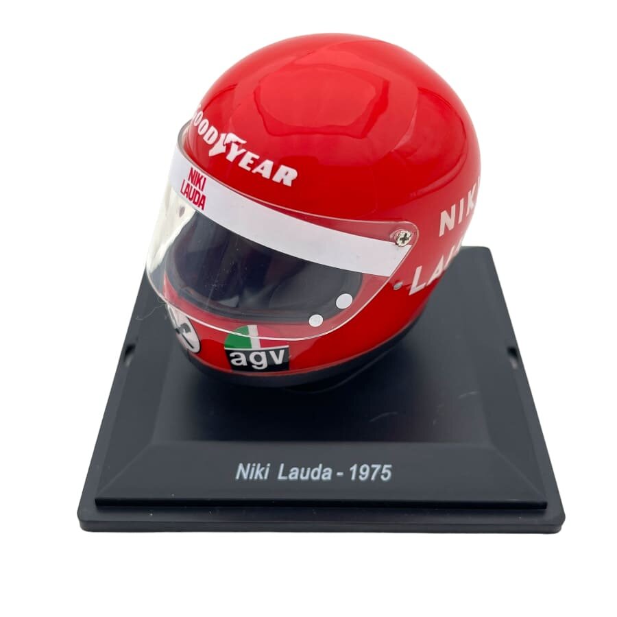 Czerwony kask z napisem \'Niki Lauda\' i logo \'Goodyear\' oraz \'agv\', umieszczony na czarnej podstawce z napisem \'Niki Lauda - 1975\'.