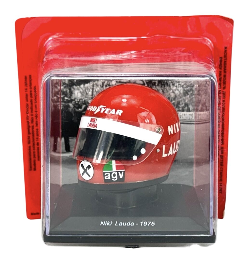 Czerwony kask z napisem \'NIKI LAUDA\' i logo \'GOODYEAR\' oraz \'agv\' umieszczony w przezroczystej gablotce. Na podstawie widnieje napis \'Niki Lauda - 1975\'.