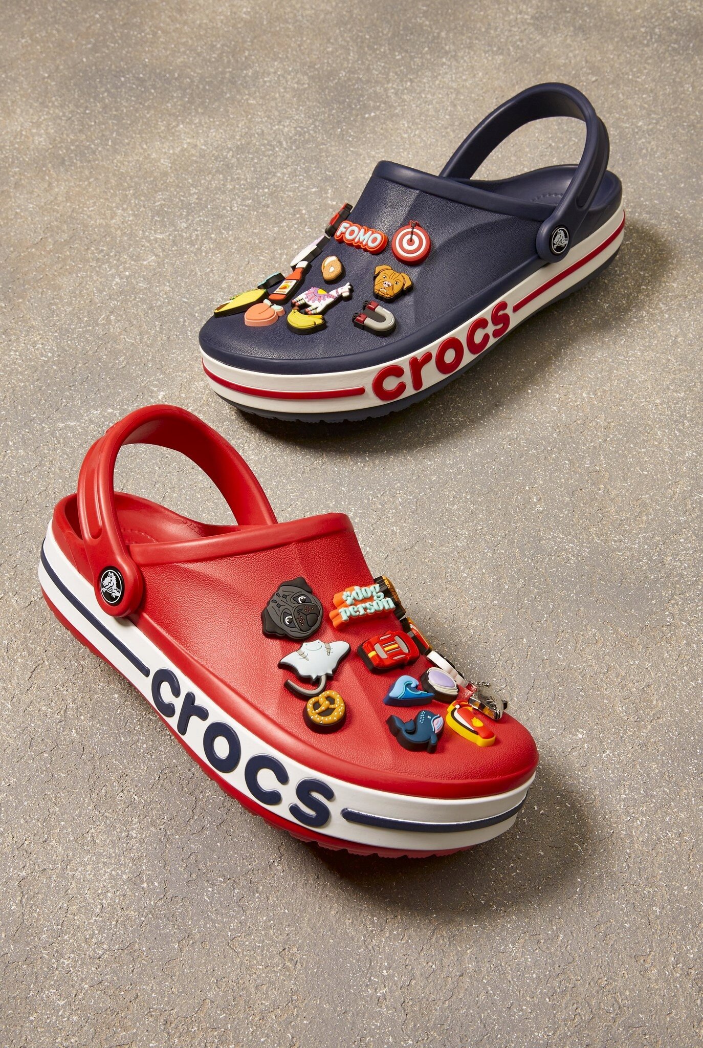Na obrazie widoczne są dwa chodaki Crocs, jeden czerwony, drugi granatowy, ozdobione różnorodnymi przypinkami. Na bokach obu butów znajduje się napis \'crocs\'.