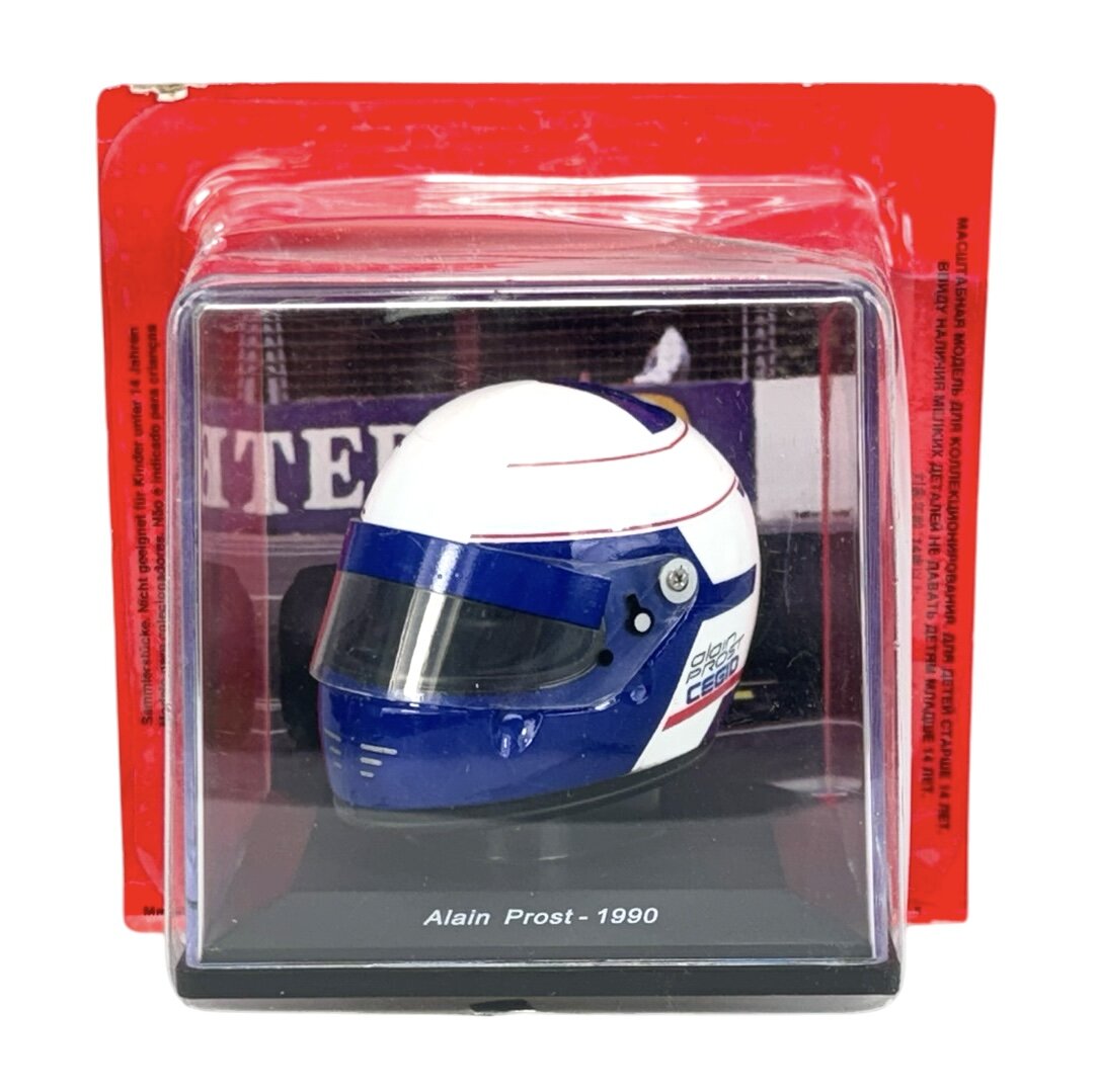 Miniaturowy kask wyścigowy w kolorach białym i niebieskim, umieszczony w przezroczystej gablotce z napisem \'Alain Prost - 1990\'.