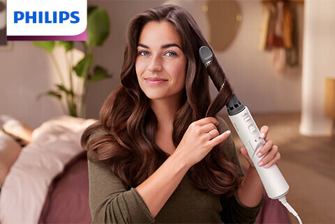 Osoba używa suszarko-lokówki do stylizacji włosów w domowym wnętrzu. W lewym górnym rogu widoczne jest logo PHILIPS.