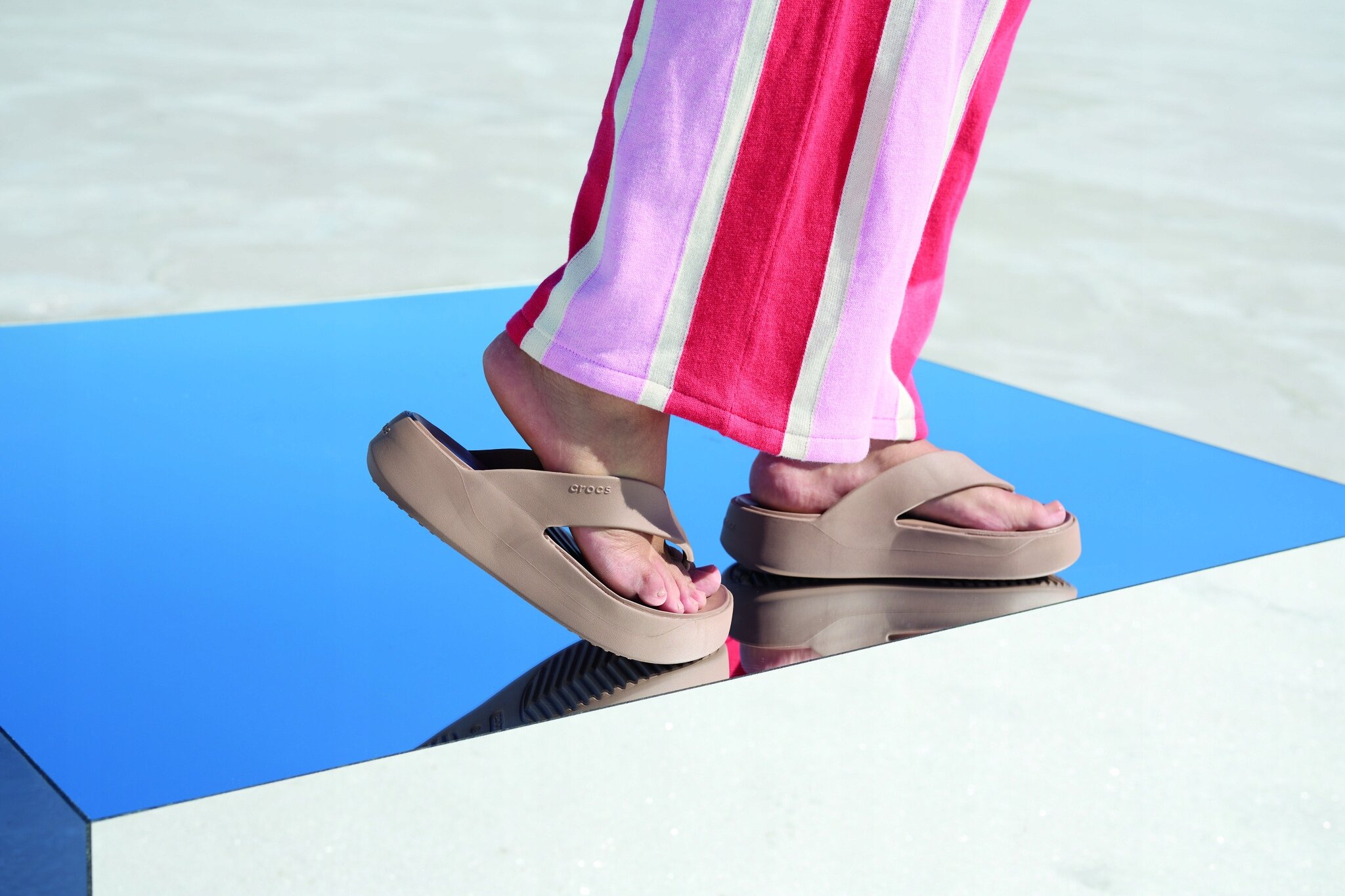 Osoba stoi na lustrzanej powierzchni, nosząc beżowe klapki z logo \'crocs\'. Ubrana jest w spodnie w pionowe paski w kolorach różowym, czerwonym i białym.