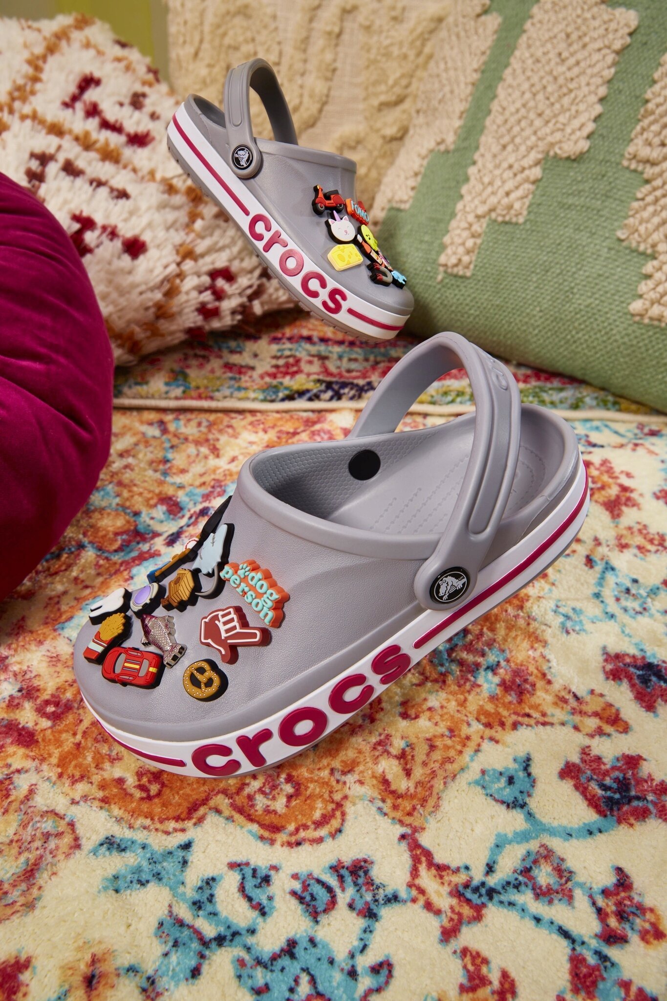 Szare chodaki z napisem \'crocs\' na boku, ozdobione kolorowymi przypinkami, leżą na wzorzystym dywanie obok poduszek.