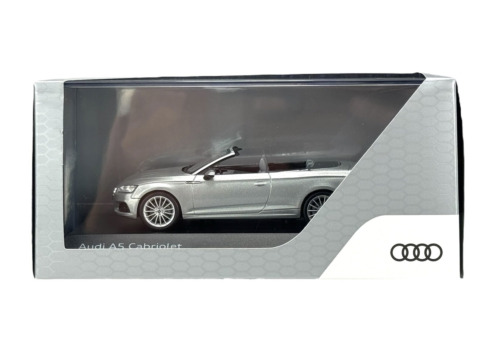 Model samochodu Audi A5 Cabriolet w kolorze srebrnym znajduje się w przezroczystym opakowaniu. Na opakowaniu widoczne jest logo Audi oraz napis \'Audi A5 Cabriolet\'.